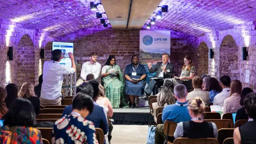A photo from the back of event, with speakers on stage and seated watchers, taking place in an underground room with exposed brickwork.